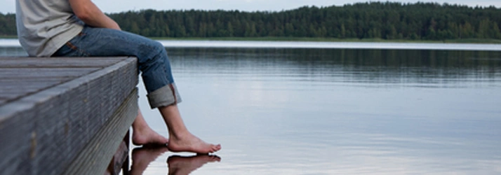 Chiropractic Wasilla AK Conditions We see a large lake with a wooden deck and a person sitting on it with their bare feet touching the water.